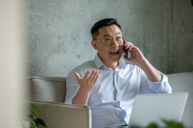 Mutlu girişimci adam kanepede oturuyor dizüstü bilgisayarda çalışıyor ve cep telefonuyla konuşuyor..