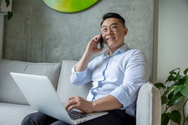 Mutlu girişimci adam kanepede oturuyor dizüstü bilgisayarda çalışıyor ve cep telefonuyla konuşuyor..