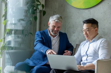 İki iş adamı modern ofiste kanepede oturmuş laptoplarını kullanıyorlar..