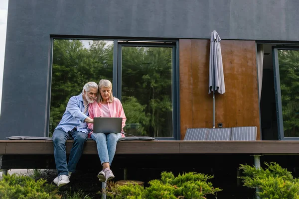 Mature couple using laptop in front of solar panels at home.