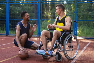 Dışardaki sahada tekerlekli sandalyede sakat bir arkadaşla basketbol oynayan, oyun oynarken konuşan bir adam..