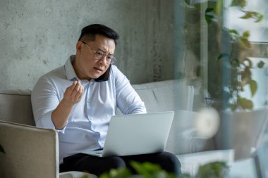 Sinirli, stresli bir girişimci kanepede oturuyor dizüstü bilgisayarda çalışıyor ve cep telefonuyla konuşuyor..