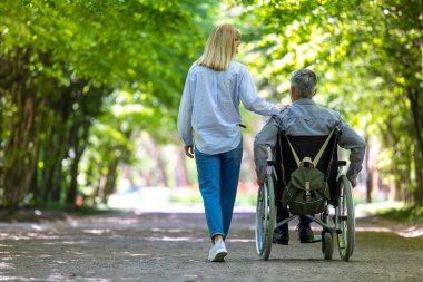 Tekerlekli sandalyedeki engelli adam karısıyla parkta yürüyor..