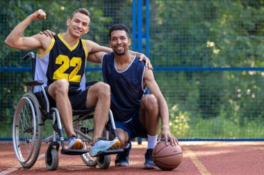 Tekerlekli sandalyedeki beyaz adam, dışarıda arkadaşlarıyla basketbol oynuyor. Yumruğunu sıktı ve gücünü gösterdi..