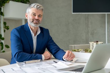 Gri saçlı, sakallı bir mimar. Bir proje üzerinde çalışıyor. Masasında dizüstü bilgisayarının önünde oturuyor..