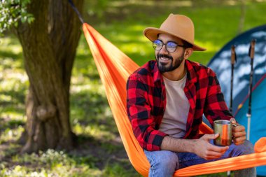 Doğanın tadını çıkarıyorum. Elinde fincanla gülümseyen adam hamakta doğanın tadını çıkarıyor.