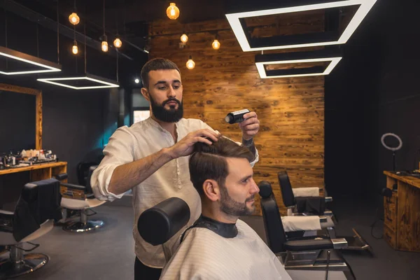 Bearded barber makes making hair treatment procedures for client in barbershop.