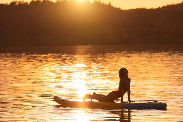 Beautiful woman practicing yoga on paddle sup surfboard at sunset.
