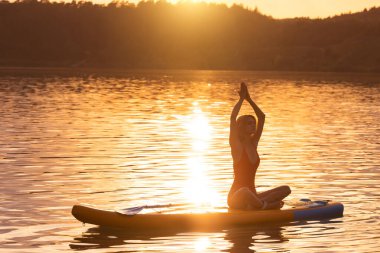 Yoga pozisyonunda genç bir kadın gündoğumunda ya da günbatımında sakin denizde sörf tahtası üzerinde pratik yapıyor..