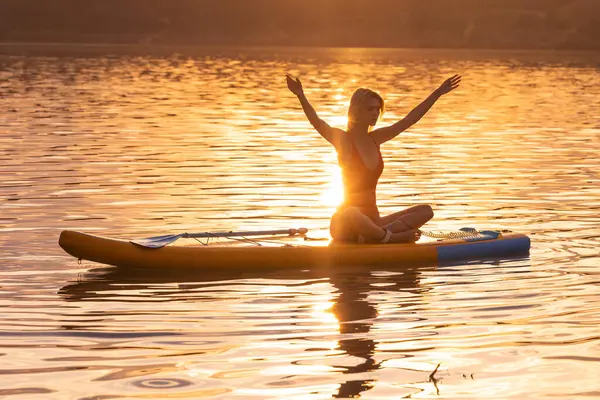 Beautiful woman practicing yoga on paddle sup surfboard at sunset.
