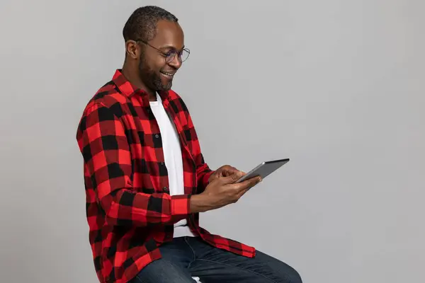 Happy black bearded man wearing red checkered shirt and glasses using digital tablet posing isolated over gray studio background.