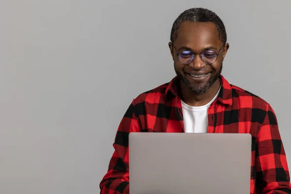 穿着红色格子衬衫的黑胡子男人微笑着在笔记本电脑上工作 在灰色工作室背景下摆出一副孤立的样子 — 图库照片
