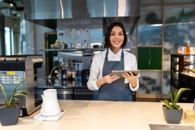 Kahve dükkanı. Elinde tabletle gülümseyen hoş, koyu renk saçlı bir kahve dükkanı asistanı.