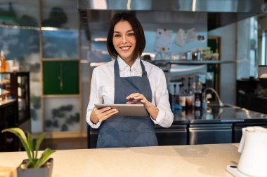 Kahve dükkanı. Elinde tabletle gülümseyen hoş, koyu renk saçlı bir kahve dükkanı asistanı.