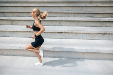 Egzersiz yapıyorum. Siyah spor giysili sarışın kadın koşuyor ve konsantre görünüyor.