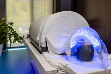 LED lambanın altında uzanan koruyucu gözlüklü kadın güzellik salonunda renkli ışık terapisi görüyor..