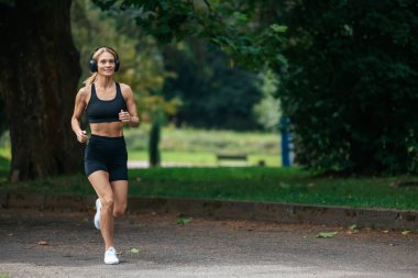 Doğal zindelik. Yaşam tarzı hareket halinde. Açık hava maratonu. Atletik güçlü sarışın koşucu kulaklıklı kadın parkta koşuyor..