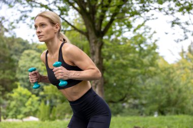 Sportif vücut. Temiz hava egzersizi. Ağırlık kaldırma. Atletik kadın dambıllarla antrenman yapıyor. Parkta çalışıyor..