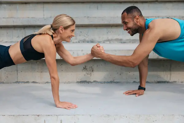 Healthy lifestyle. Exercise support. Multiracial sportive woman and man holding hands while standing in plank outdoor.