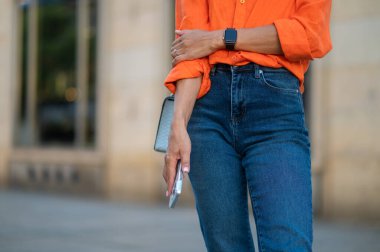 Kot pantolonlu tanınmayan bir kadın dışarıda elinde akıllı telefonuyla dikiliyor..