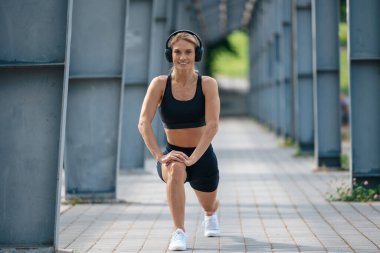 Gülümseyen fitness kadını kulaklık takıyor, spor yapıyor ve dışarıda bacaklarını açıyor..