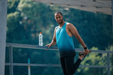 Bacak esnetiyorum. Koyu renk saçlı, spor kıyafetli, egzersiz yapan ve bacaklarını esneten bir adam.