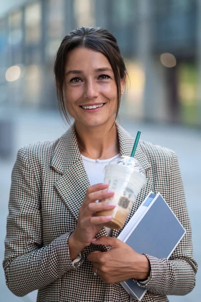 Smiling confident woman wearing official style clothing posing outdoor drinking milk cocktail and holding organizer.