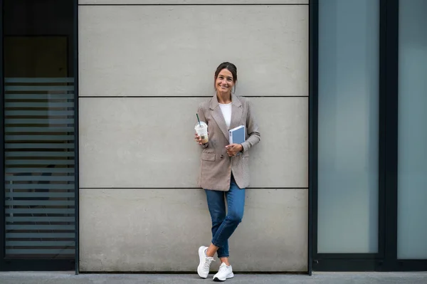 Stylish confident woman with cocktail and notebook standing near modern building.