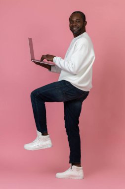 Beyaz kazak giyen neşeli siyah adam pembe arka planda uzaktan izole edilmiş bilgisayarla çalışıyor..