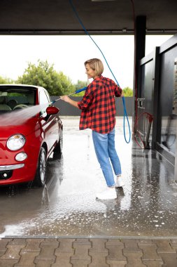 Araba yıkama. Genç sarışın kadın arabayı hortumla yıkıyor.