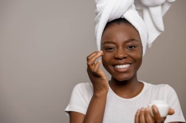 Kafasında havlu olan neşeli çekici kadın günlük cilt bakımını yapıyor krem nemlendirici ile kahverengi arka planda izole edilmiş.