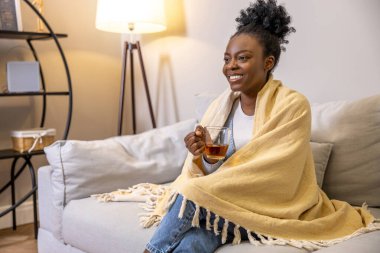 Çekici, gülümseyen bir kadın, kanepede otururken çay içiyor. Evin içinde sarı bir ekoseye sarılı.