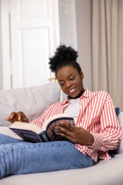 Gülümseyen Afrikalı Amerikalı kadın evde kanepede otururken kitap okuyor..