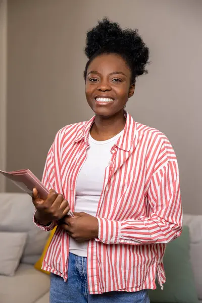 Happy woman real estate agent with afro hairstyle holding folder and smiling at camera in new house.
