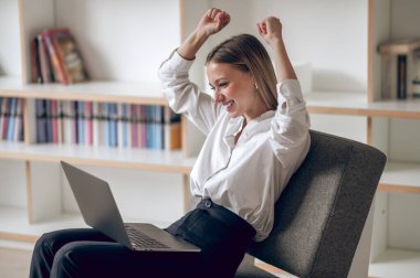 Oturma odasında otururken dizüstü bilgisayar kullanan neşeli beyaz kadın yumruklarını sıkarak seviniyor..