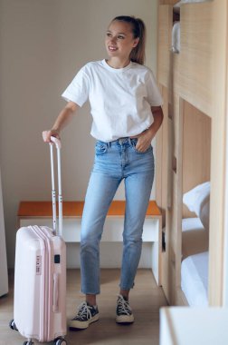 Beyaz gömlekli ve kot pantolonlu kendine güvenen bir kadın otel odasında bavuluyla duruyor..