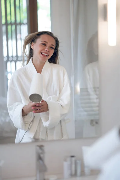 Woman with hair dryer. Joyful woman in white robe with a hair dryer feeling happy