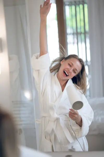 Woman with hair dryer. Joyful woman in white robe with a hair dryer feeling happy