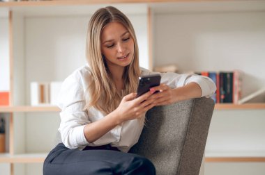 Beyaz tişörtlü, mutlu, güzel bir kadın. Kıyafetinin içinde otururken cep telefonu kullanıyor..