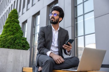 Koyu renk saçlı Hindu işadamı dışarıda çalışıyor ve telefonla konuşuyor.