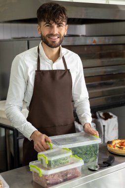Koyu saçlı, sakallı, genç pizzacı konteynırlara yiyecek dolduruyor.