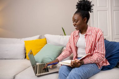 Afro saçlı güzel bir kadın panterine notlar yazıyor ve oturma odasındaki koltukta dizüstü bilgisayar kullanıyor..