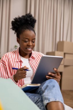 Afro saç stili yaratıcı kadın rahat bir koltukta oturup tablet üzerinde çalışırken günlüğüne not alıyor..