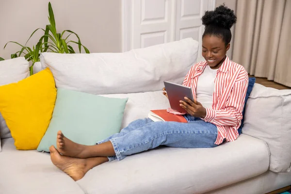 Woman with afro hairstyle holding tablet pc browsing web pages while relaxing on sofa at home.