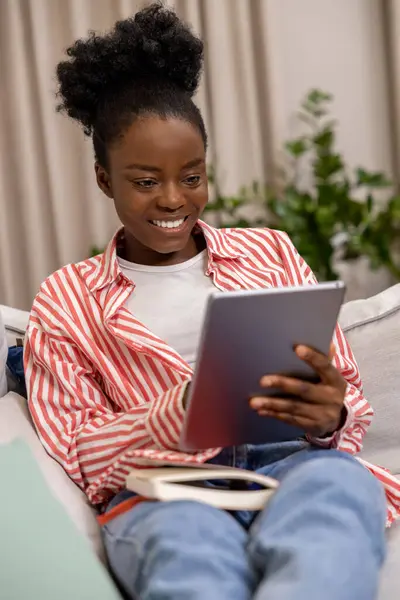 Smiling black woman using digital tablet for checking social media sitting on couch in living room.