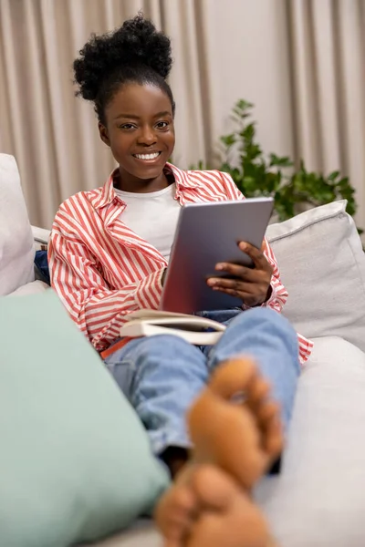 Woman with afro hairstyle holding tablet pc browsing web pages while relaxing on sofa at home.