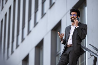 Esmer Hintli genç adam, binanın arka planında, elinde telefonla.