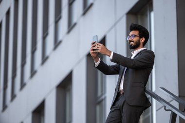 Esmer Hintli genç adam, binanın arka planında, elinde telefonla.