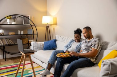 Neşeli kadın ve erkek akşam yemeği için pizza yiyorlar bilgisayardan film izliyorlar evdeki koltukta oturuyorlar