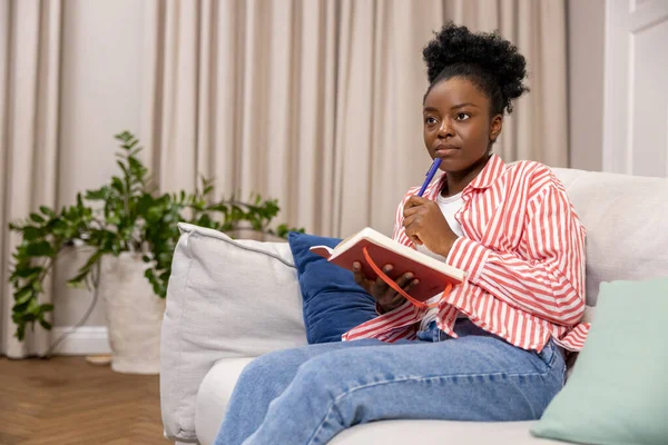Black woman writing to do list or plan for day while sitting on couch in home interior.